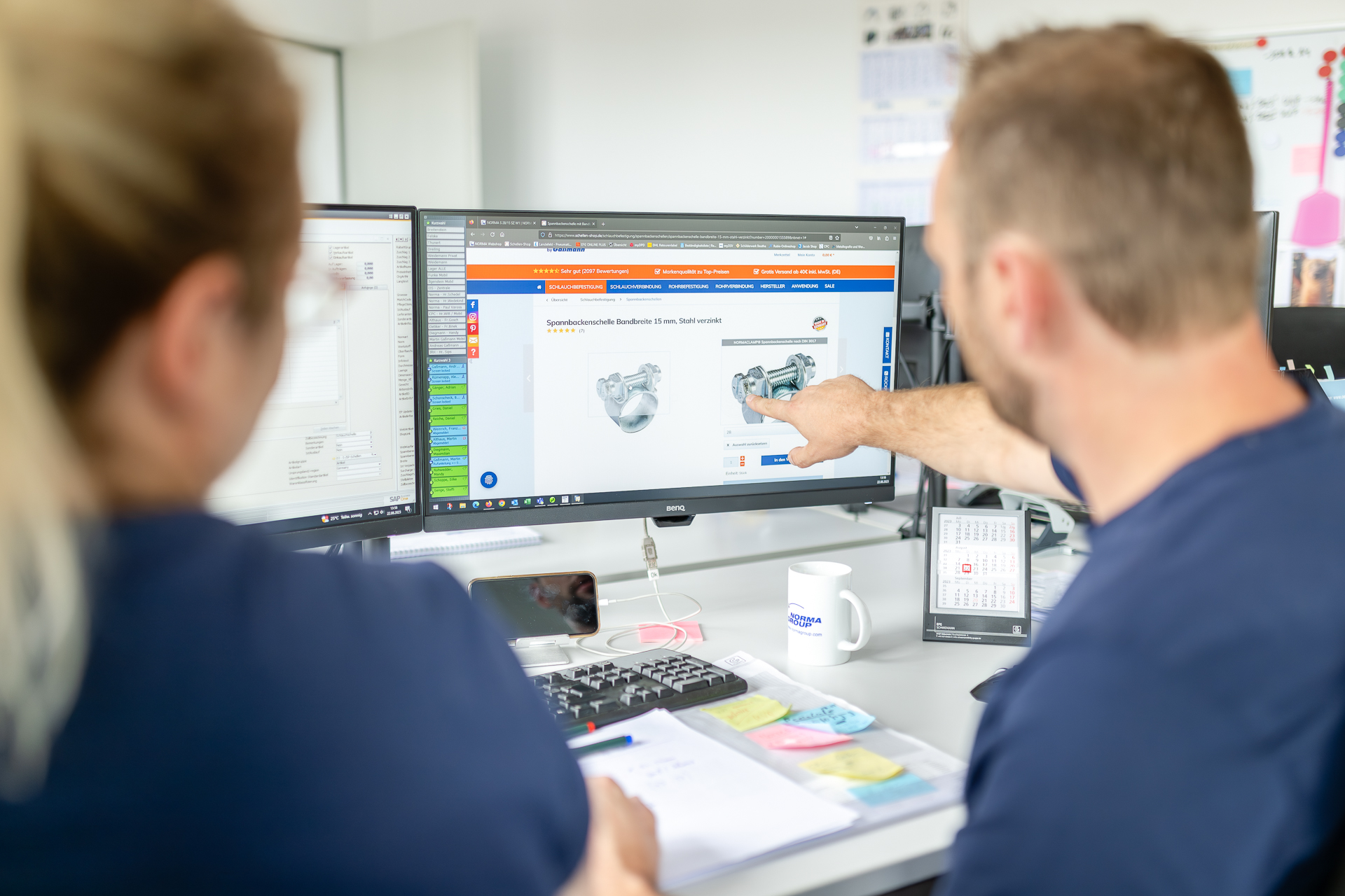 Mitarbeiter analysieren eine Spannbackenschelle auf einem Produktbildschirm. Zwei Personen betrachten am Computerbildschirm eine verzinkte Spannbackenschelle, wobei eine Person mit dem Finger darauf zeigt.
