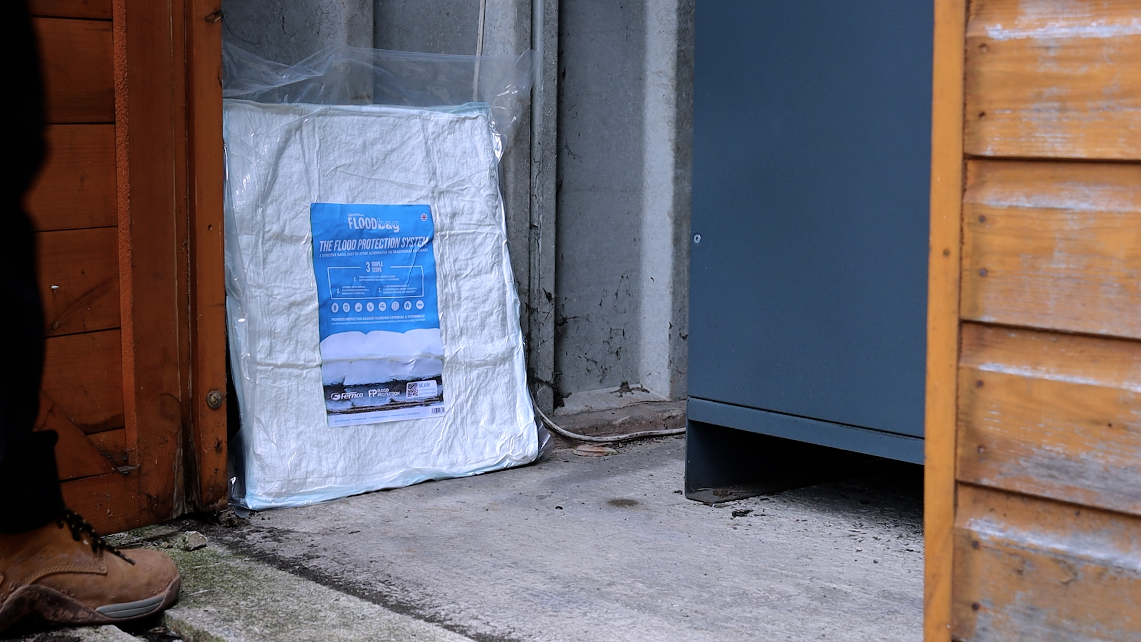 Ein in Folie verpackter Fernco FLOODbag, ein moderner Sandsack für Hochwasserschutz, liegt auf dem Boden vor einer Türöffnung. Das blaue Produktetikett mit der Aufschrift 'FLOODbag' ist deutlich sichtbar.