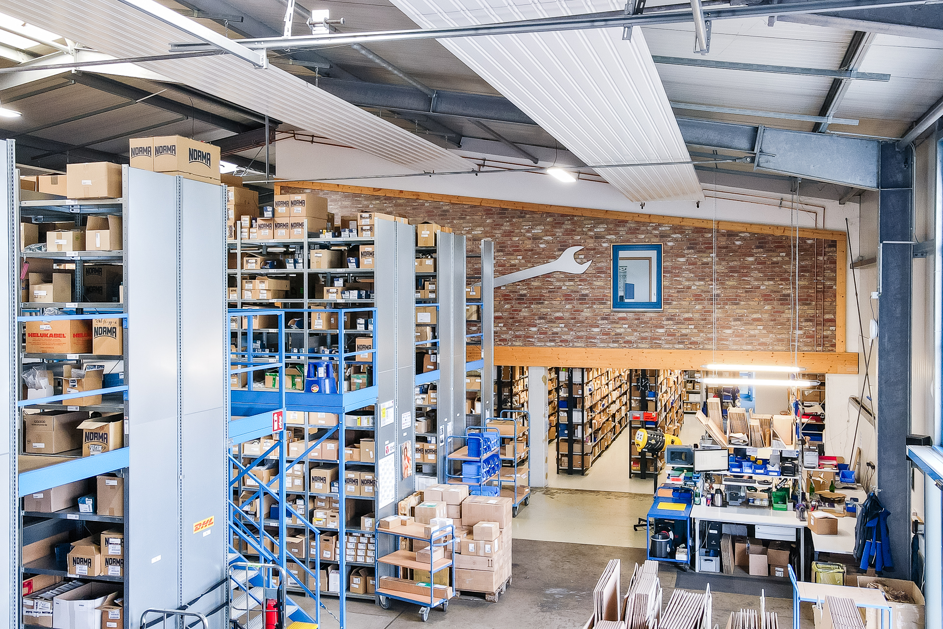 Modernes Lager- und Logistikzentrum mit Regalen und Arbeitsbereich Große Lagerhalle mit hohen Regalsystemen, gefüllt mit Kartons und Produkten. Links und mittig stehen graue und blaue Regale, teils mehrstöckig. Im Hintergrund eine Backsteinwand mit einem großen weißen Schraubenschlüssel und einem blauen Fenster über eine