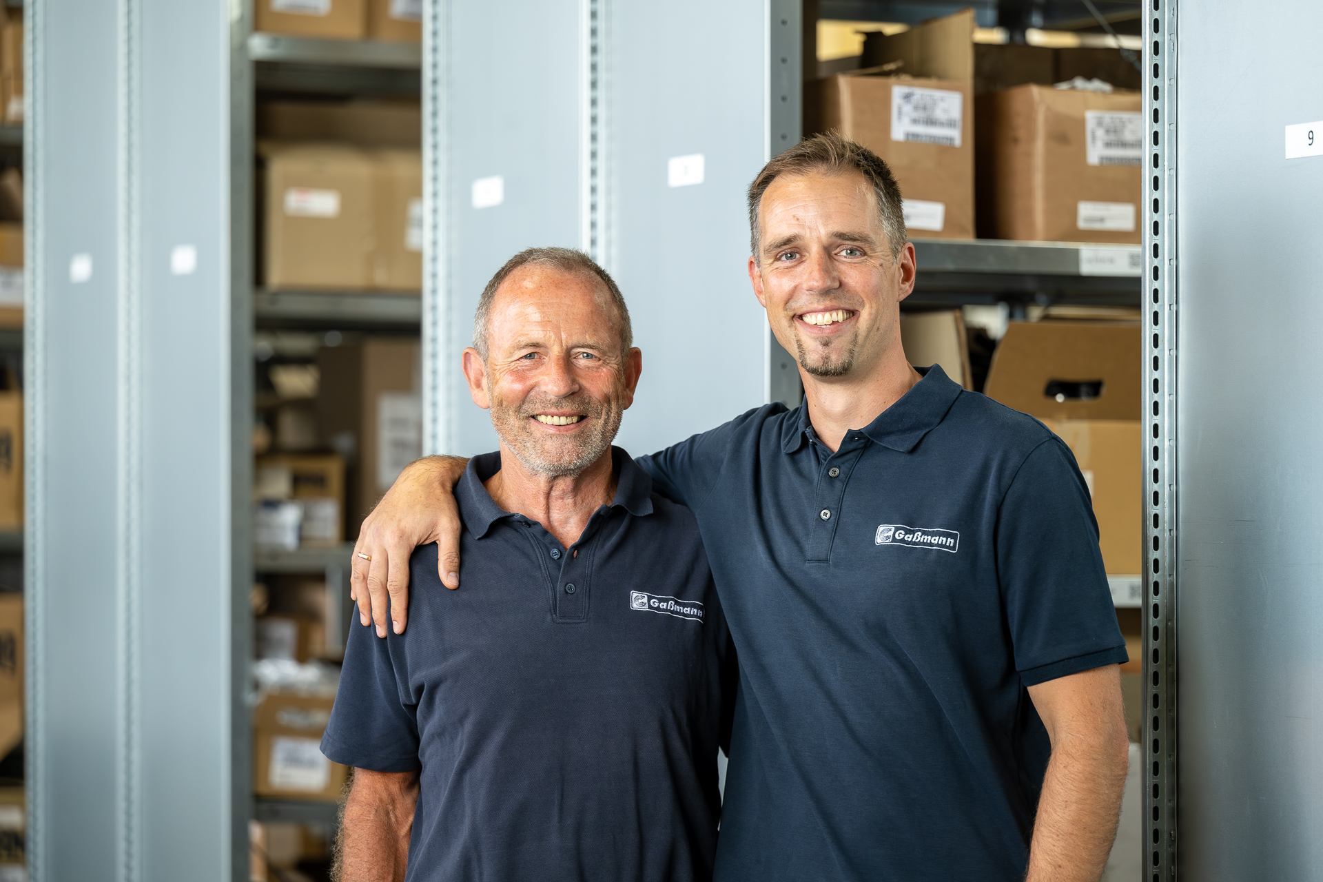 Geschäftsführung der Gaßmann GmbH – Martin und Andreas Gaßmann Zwei lächelnde Männer in dunkelblauen Gaßmann-Poloshirts stehen eng beieinander in einem Lager, dessen Regale mit Kartons gefüllt sind.