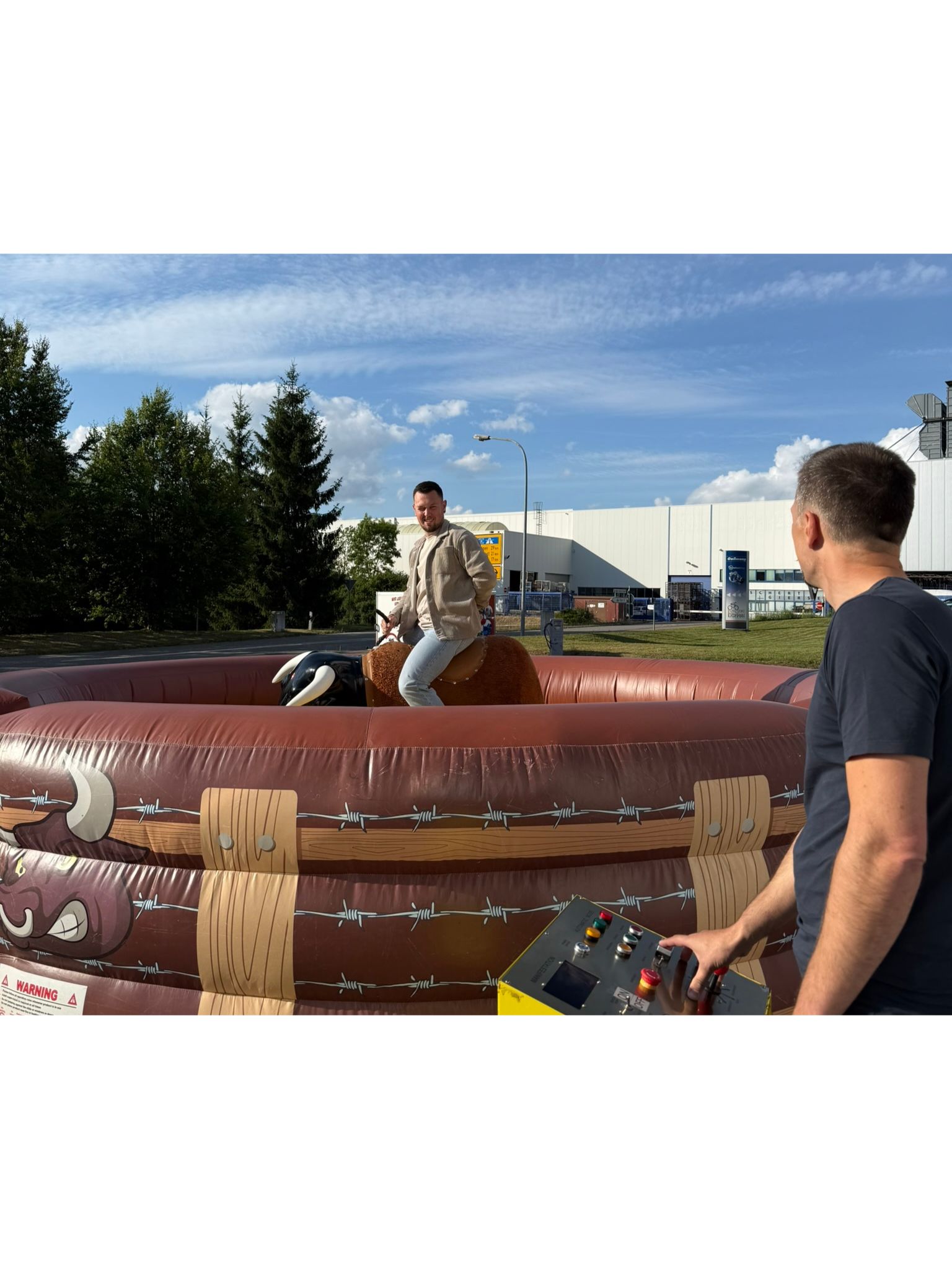 Ein Teilnehmer des Sommerfests 2026 der Gaßmann GmbH beweist seinen 'festen Halt' beim mechanischen Bullenreiten, während ein Mitarbeiter das Gerät bedient. Eine fröhliche Teambuilding-Aktion im Freien unter blauem Himmel.