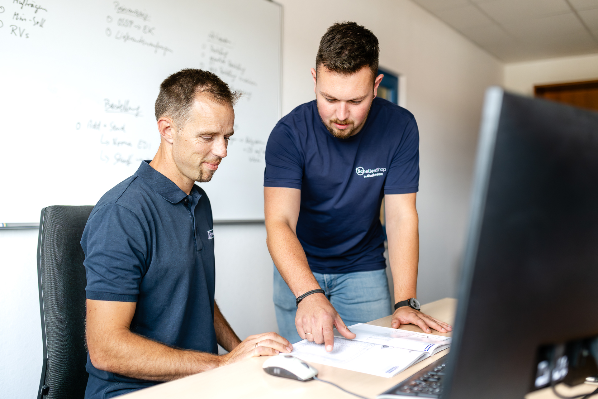 Mitarbeiter von SchellenShop bei der gemeinsamen Dokumentenprüfung am Schreibtisch. Zwei Mitarbeiter in dunklen Shirts besprechen Dokumente an einem Schreibtisch. Der stehende Mitarbeiter zeigt auf Unterlagen, während der sitzende aufmerksam zuschaut. Im Hintergrund ist eine beschriebene Whiteboard-Tafel zu sehen.