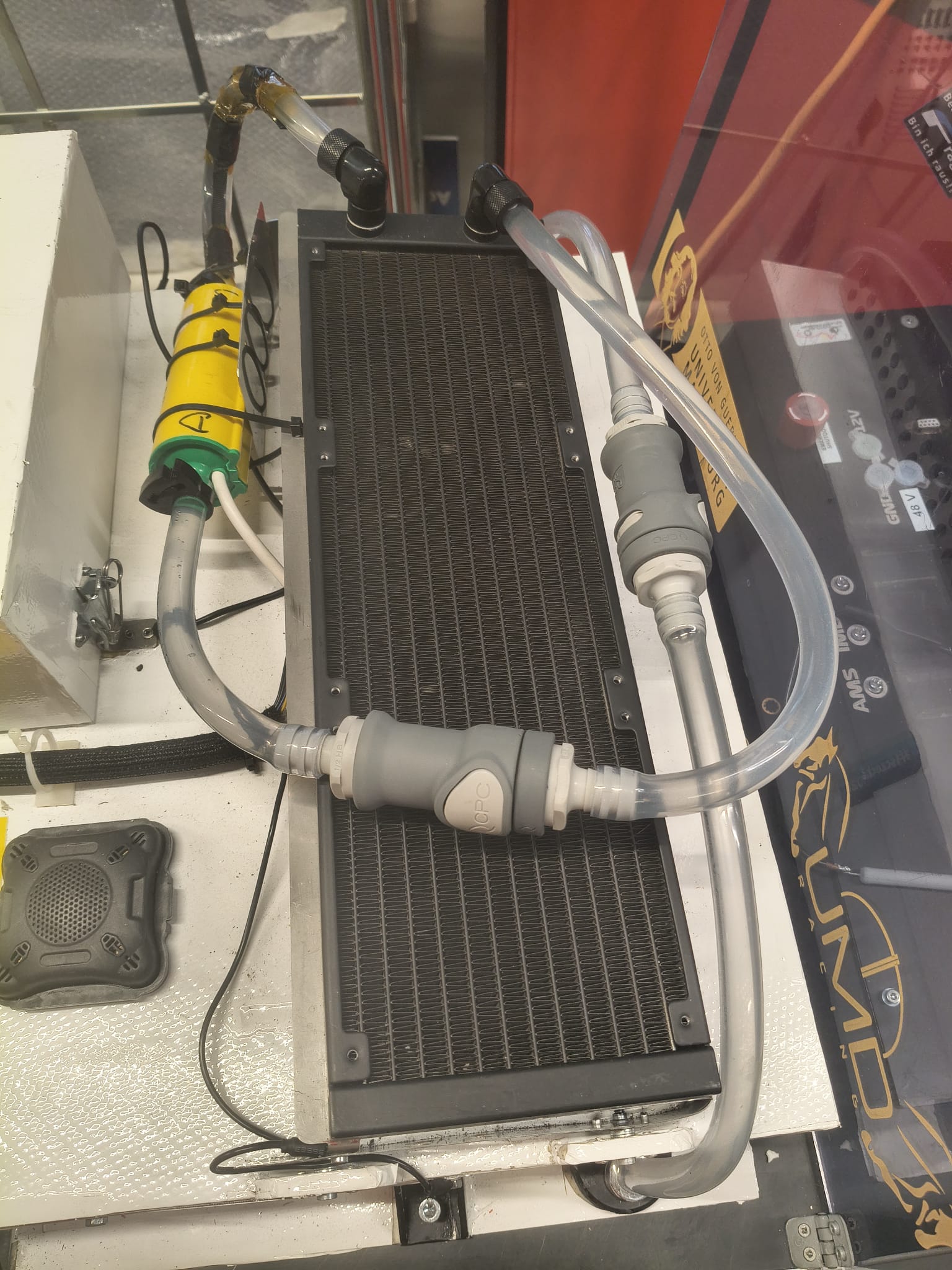 Detailansicht eines Akkukühlsystems mit einem schwarzen Kühler, transparenten Schläuchen und grauen CPC Schnellkupplungen sowie einem gelben zylindrischen Bauteil. Dieses System wird vom Schellen-Shop für UMD Racing e.V. bereitgestellt.
