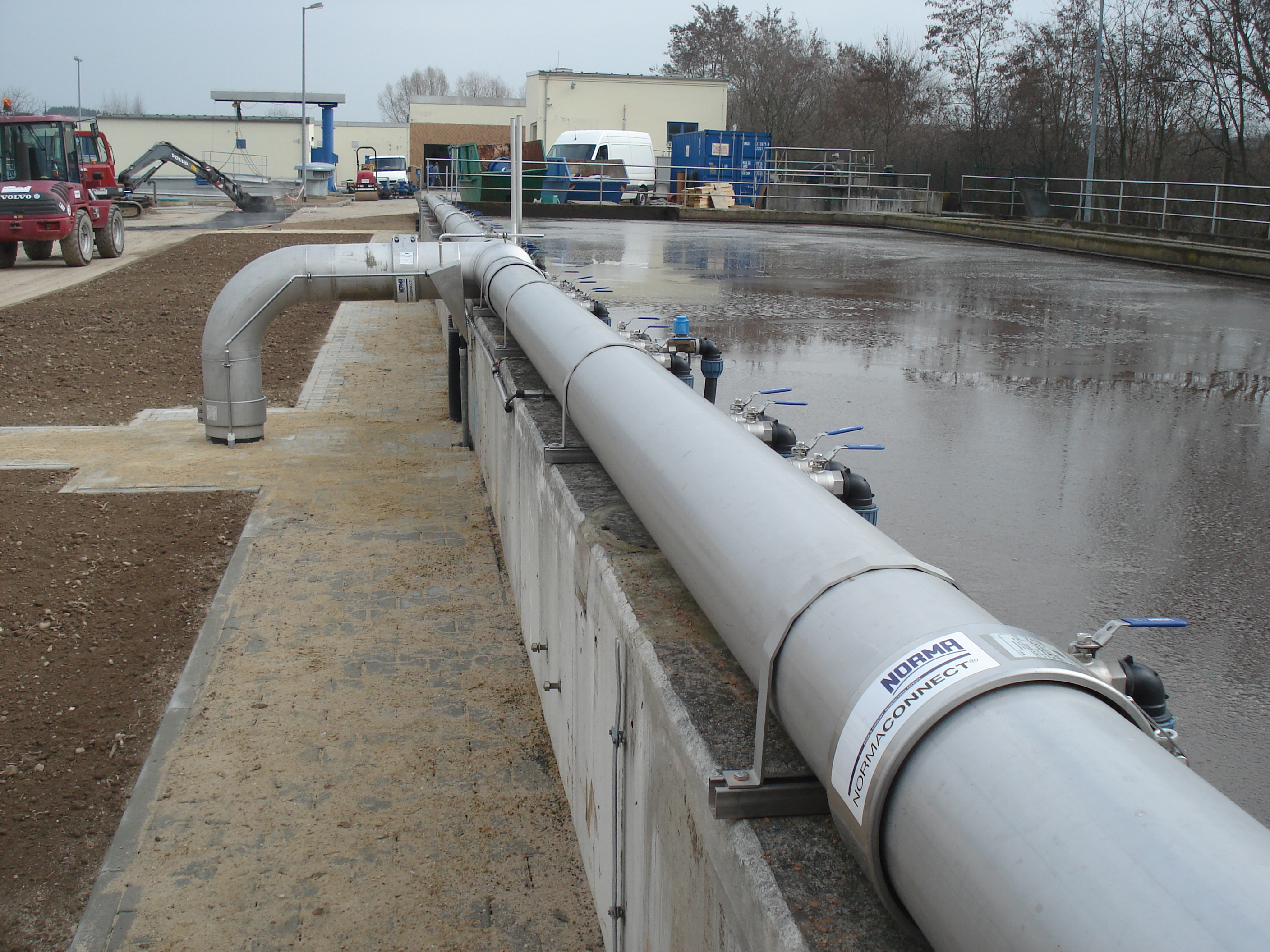 Robuste graue Rohrleitung mit NORMA CONNECT Kupplungen und blauen Ventilen, montiert an einer Betonwand, symbolisiert 'Sicher und Stark: Rohrkupplungen für hohe Drücke'.