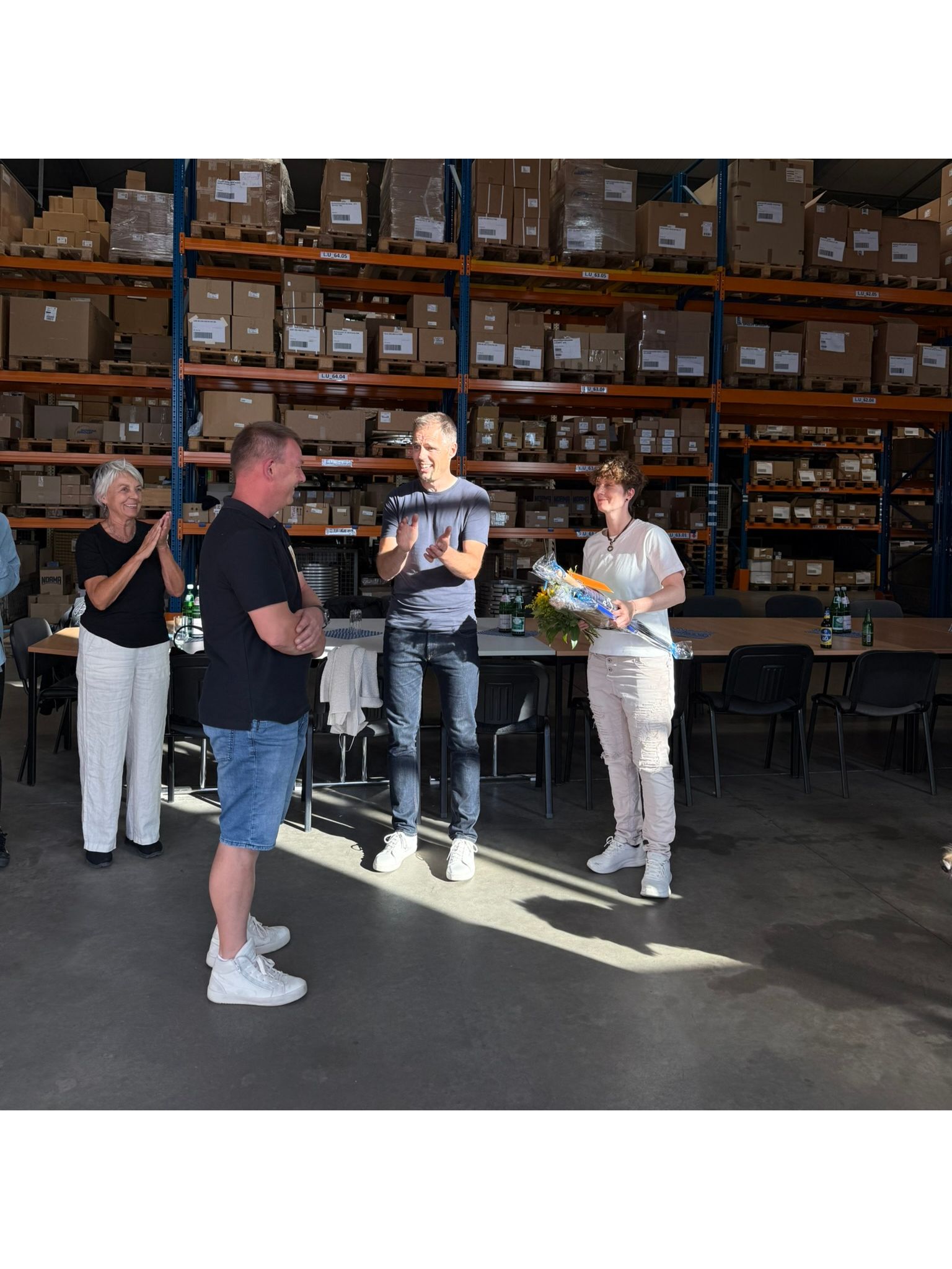 Vier lächelnde Mitarbeiter der Gaßmann GmbH feiern ihr Sommerfest 2026 in einer Lagerhalle, umgeben von Regalen voller Kartons. Eine Frau hält einen Blumenstrauß und ein verpacktes Geschenk, während Kollegen applaudieren, was das Motto 'Teambuilding mit f