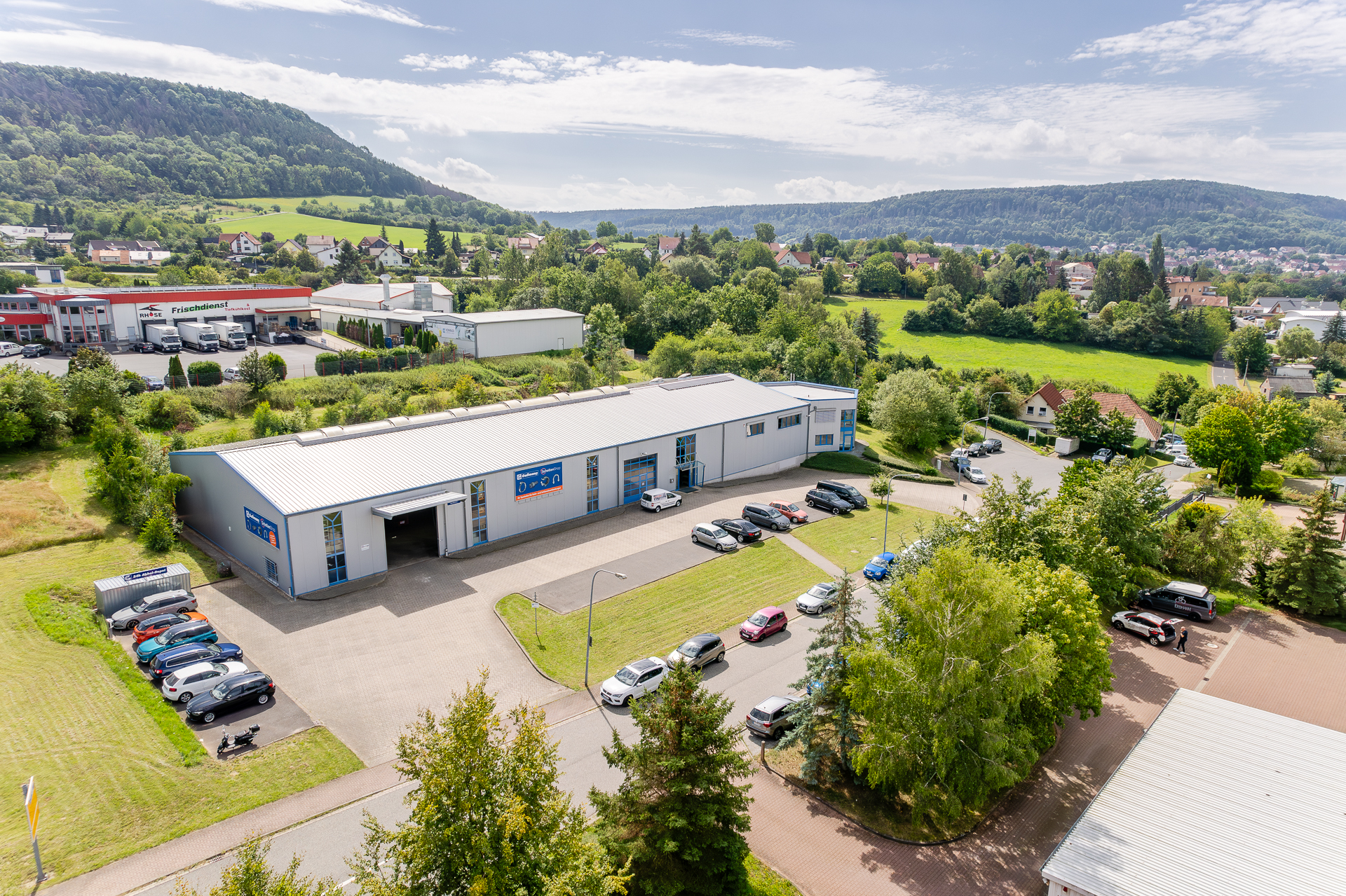 Gewerbepark mit Gaßmann GmbH und Kraftfahrzeugdienstleistungen, eingebettet in eine ländliche Landschaft unter blauem Himmel. Luftaufnahme eines Gewerbegebiets mit mehreren Firmengebäuden, darunter ein Logistikzentrum und ein KFZ-Betrieb, umgeben von grünen Hügeln und Wohnbebauung.