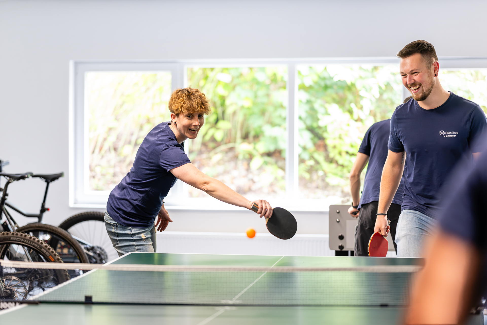 Freizeit und Teambuilding: Mitarbeiter spielen begeistert Tischtennis. Lachende Mitarbeiter spielen Tischtennis in einem modernen Raum. Eine Frau mit rotem Haar schlägt den Ball, während ein Mann im blauen SchellenShop-T-Shirt am Tisch lacht. Im Hintergrund sind Fenster und Fahrräder sichtbar.