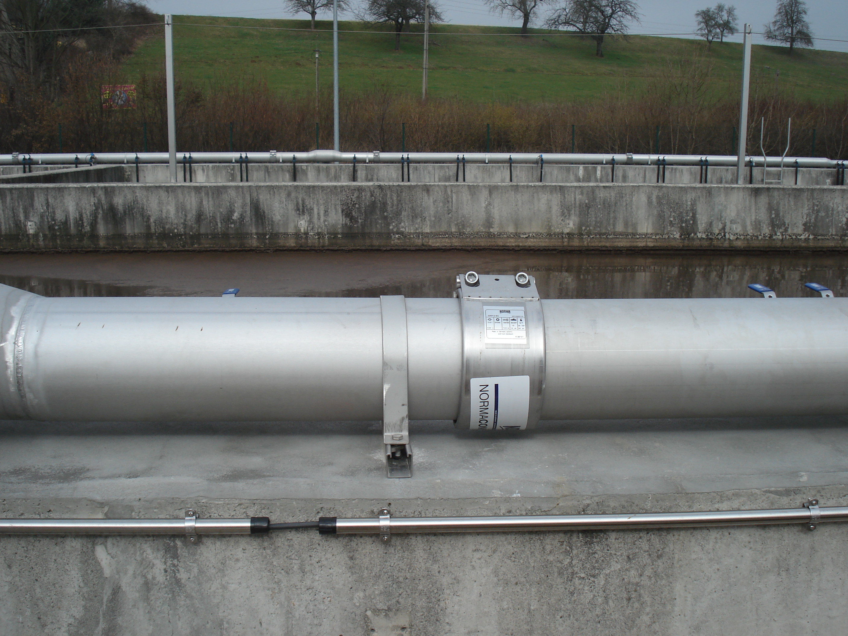 Ein großes, silberfarbenes Rohr mit einer NORMACON Hochdruck-Rohrkupplung der Marke 'Sicher und Stark', montiert auf einer Betonkonstruktion in einer Industrieumgebung. Die robuste Kupplung gewährleistet eine sichere Verbindung für hohe Drücke.