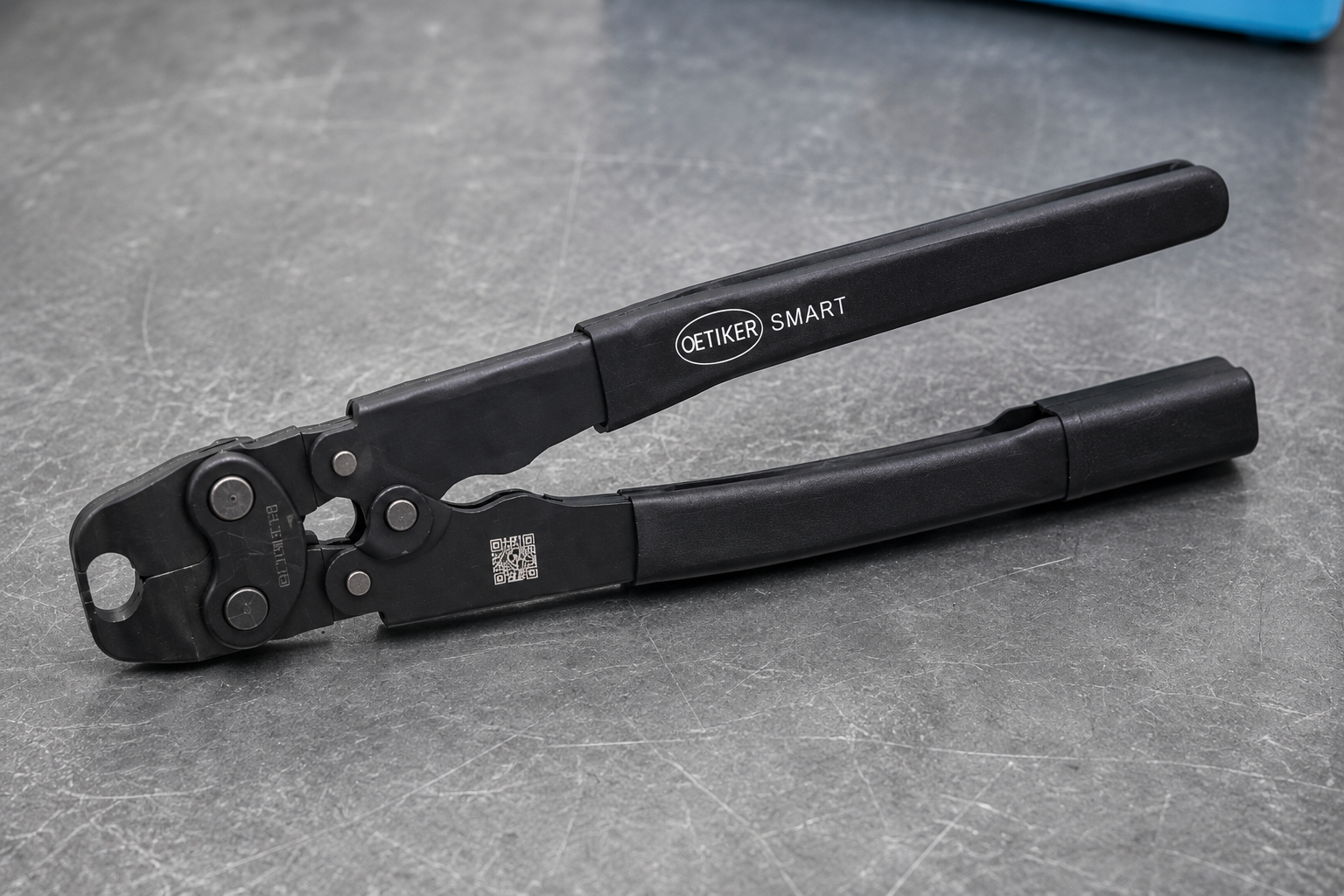 Oetiker SMART Tool, eine robuste schwarze Handpresszange mit dem Oetiker SMART Logo auf dem Griff, liegend auf einem grauen Werkstatttisch. Sie dient zum präzisen Verpressen von Schellen.