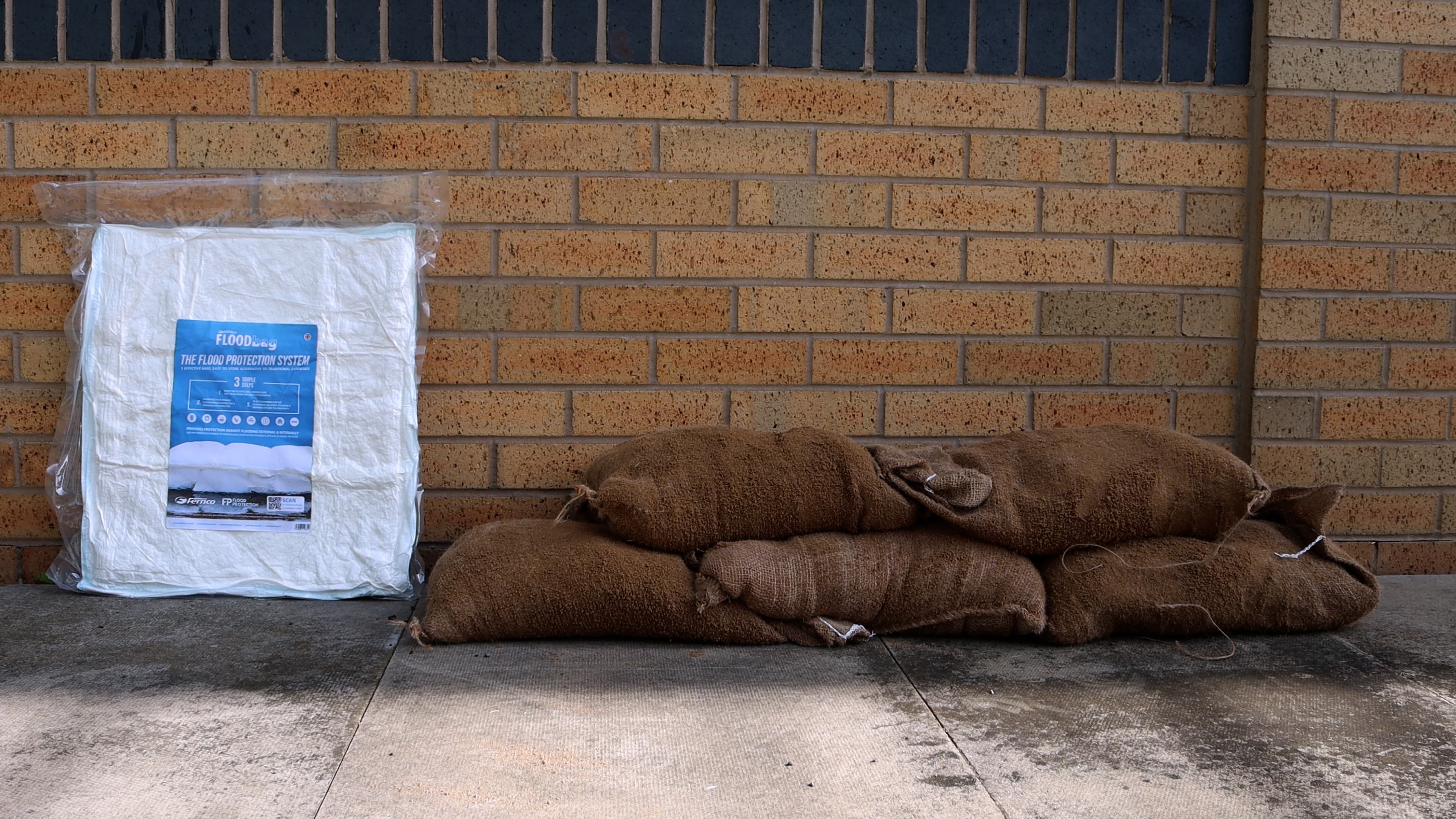 Das Fernco FLOODbag (5er Pack) – der moderne Sandsack bei Hochwasser – in transparenter Verpackung, unaktiviert und flach, steht links neben einem Stapel von drei gefüllten, braunen traditionellen Sandsäcken rechts, vor einer Ziegelwand.