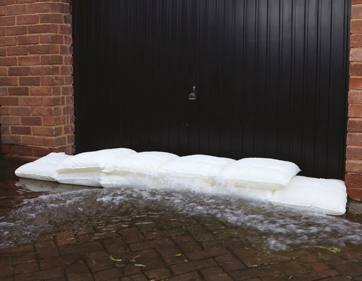 Weiße Fernco FLOODbags, moderne Sandsäcke bei Hochwasser, schützen eine schwarze Garagentür vor spritzendem Flutwasser auf einem Pflasterweg.