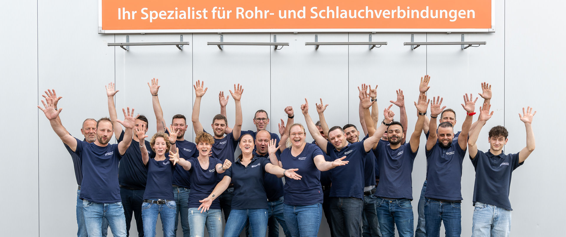 Wir sind Schellen-Shop! Mitarbeiter des Schellen-Shops stehen unter einem Banner mit dem Slogan "Ihr Spezialist für Rohr- und Schlauchverbindung"