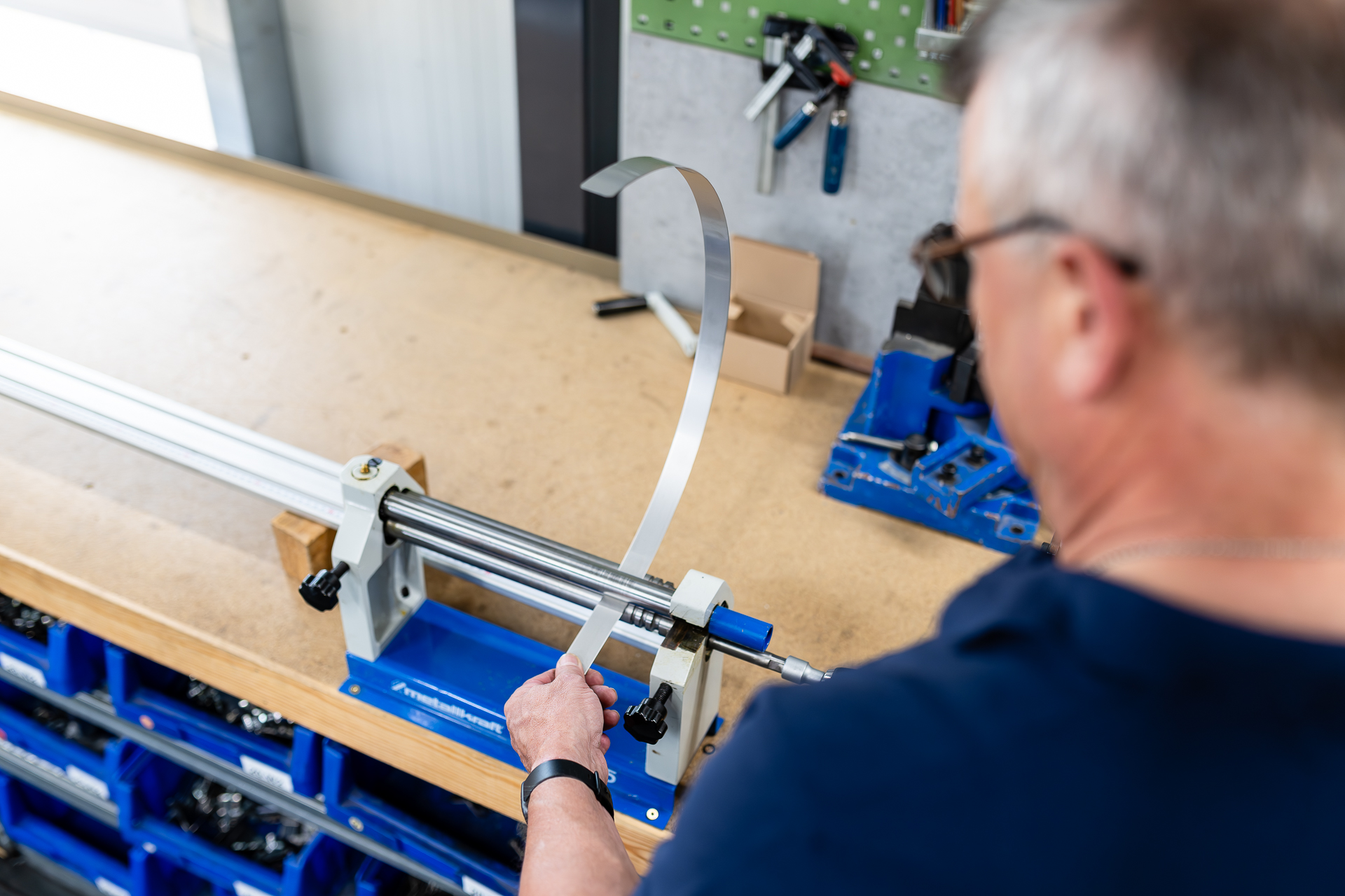 Arbeitsprozess: Präzises Biegen von Metallstreifen mit einer Handbiegemaschine. Ein Mann biegt einen langen, schmalen Metallstreifen mit einer manuellen Rollenbiegemaschine auf einer Holzwerkbank in einer Werkstatt.