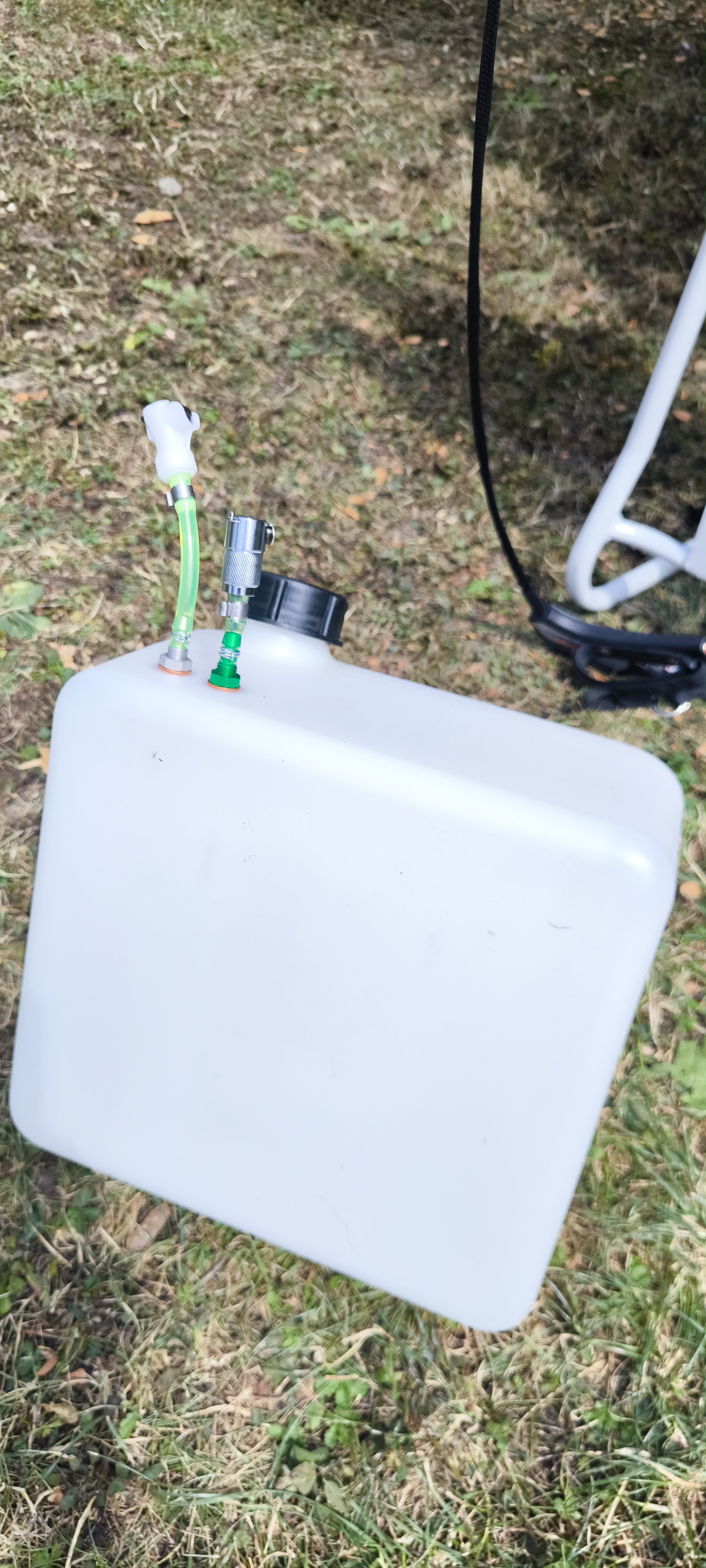 Weißer rechteckiger Kraftstofftank mit montierten grünen Benzinschläuchen und Schnellkupplungen für den Paramotor, liegend auf Gras. 'Benzinkupplungen am Paramotor – sicher tanken, sauber transportieren'.