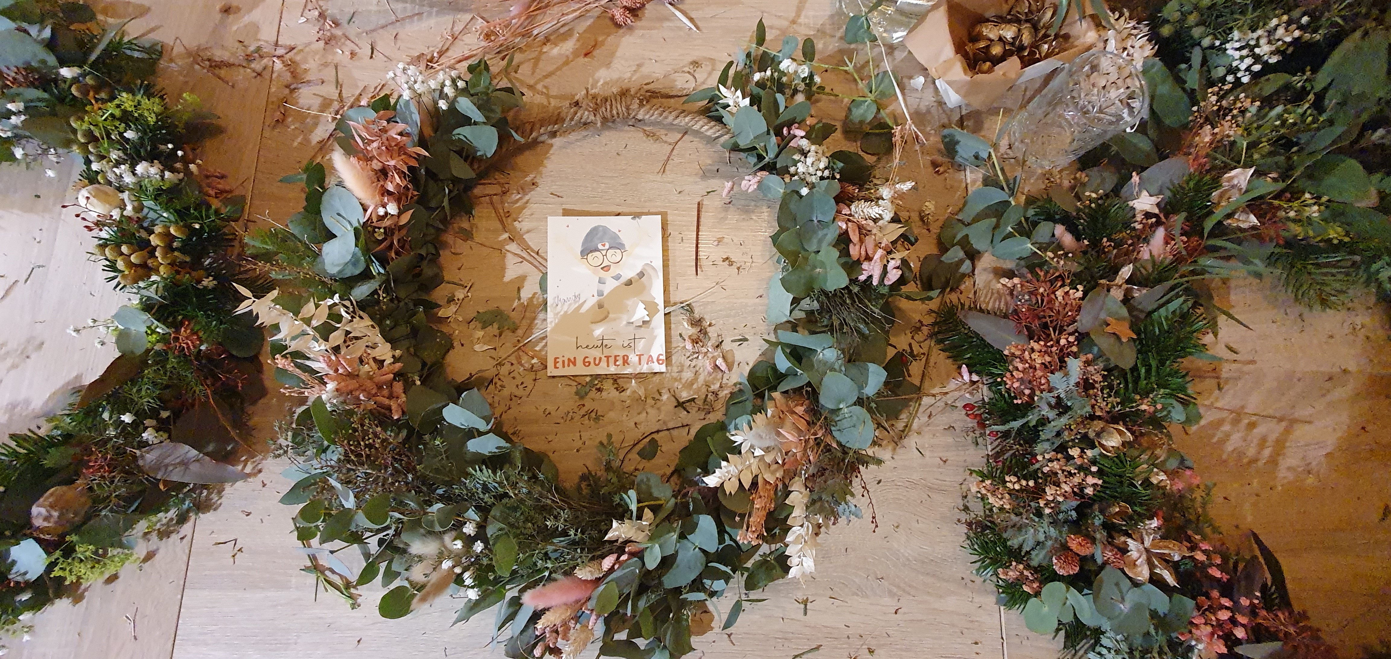 Florale Kränze und Dekorationen mit Eukalyptus, Tannenzweigen und Trockenblumen, darunter eine Karte mit der Aufschrift 'heute ist EIN GUTER TAG', erstellt während des Teambuildings 'Zauberhafte Vorweihnachtszeit bei Ly’s'.