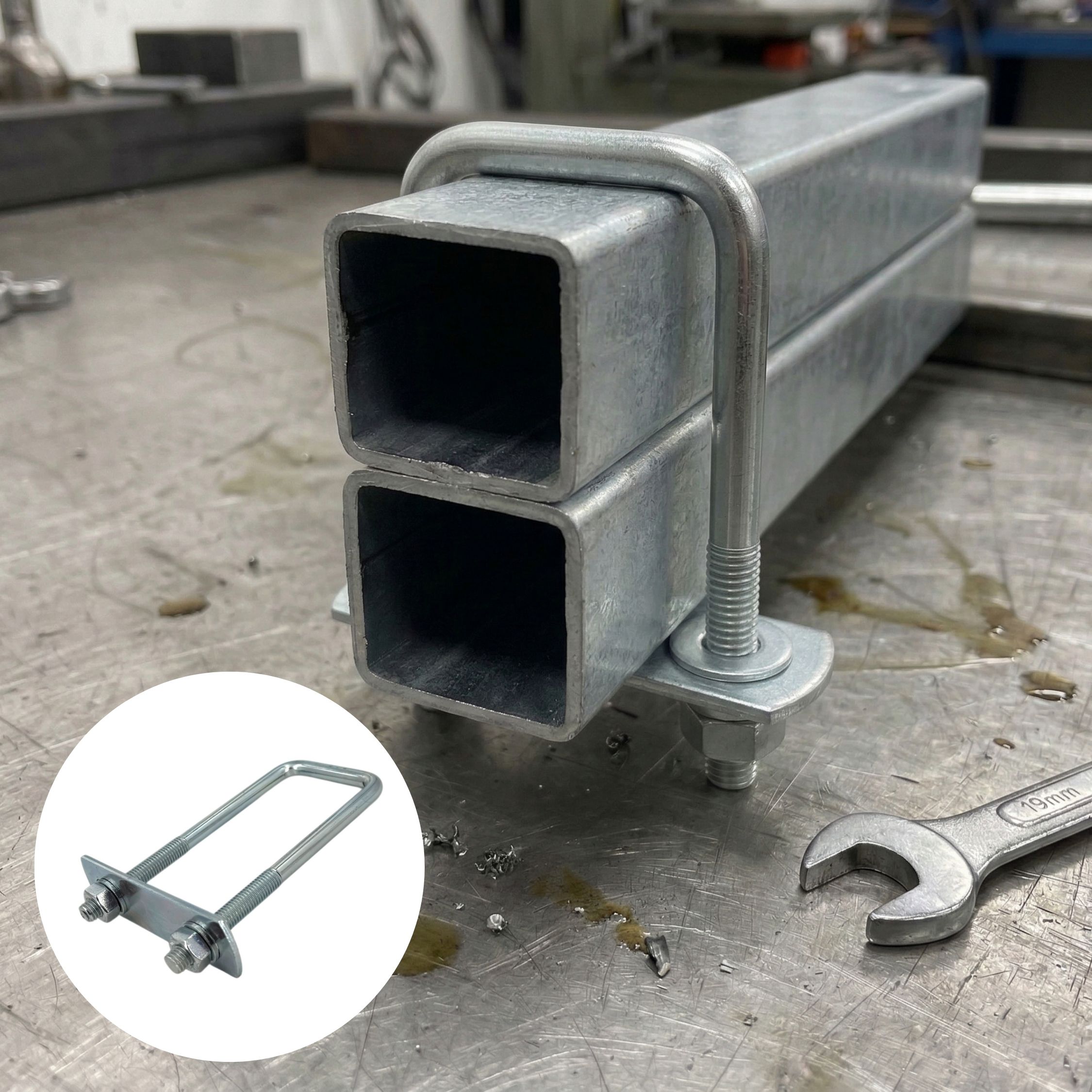 Der verzinkte Quadratrohrbügel / Rechteckbügel mit langen Schenkeln, ähnl. Typ 3575, ist auf einer Werkbank an zwei übereinanderliegenden Vierkantrohren montiert und verbindet diese fest. Im Vordergrund rechts liegt ein Schraubenschlüssel. Ein separater,