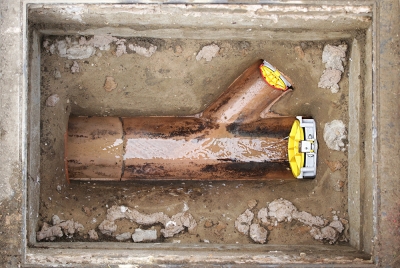 Nahaufnahme einer installierten Rückstauklappe / Froschklappe für Rohreingänge / Rohrmuffen an einem Y-förmigen Abwasserrohr in einem offenen Erdgraben.