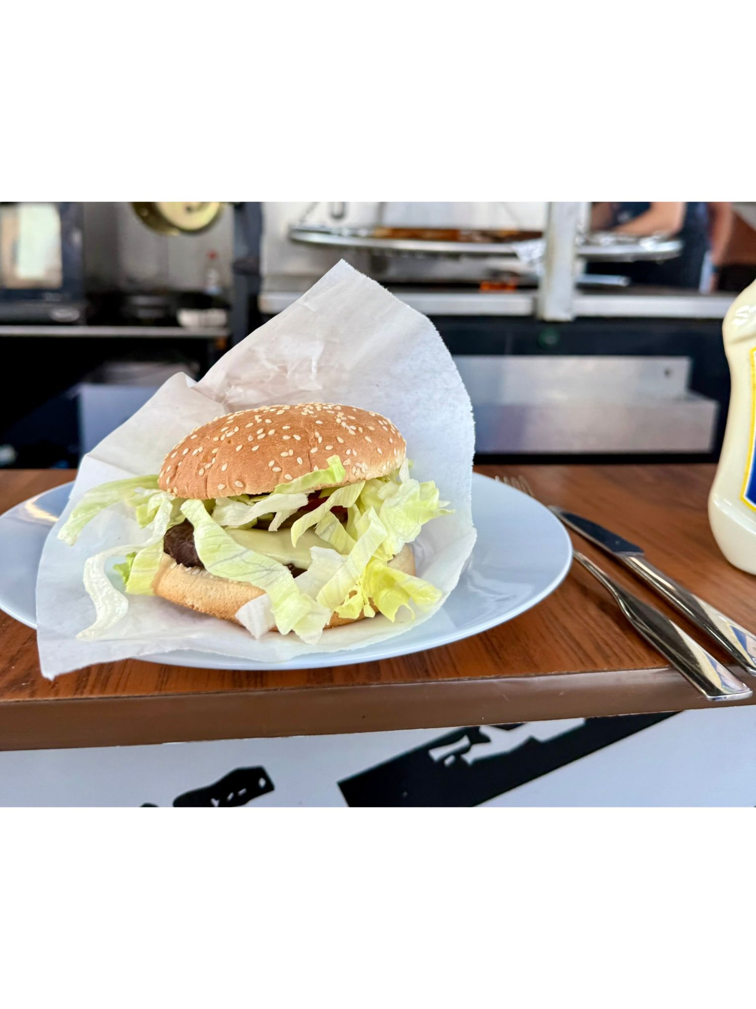 Ein appetitlicher Cheeseburger mit Sesambrötchen, saftigem Patty, geschmolzenem Käse und frischem Eisbergsalat, serviert auf einem weißen Teller vor einem Imbissstand. Dieses Bild zeigt die leckere Verpflegung beim Teambuilding mit festem Halt: Das Sommer
