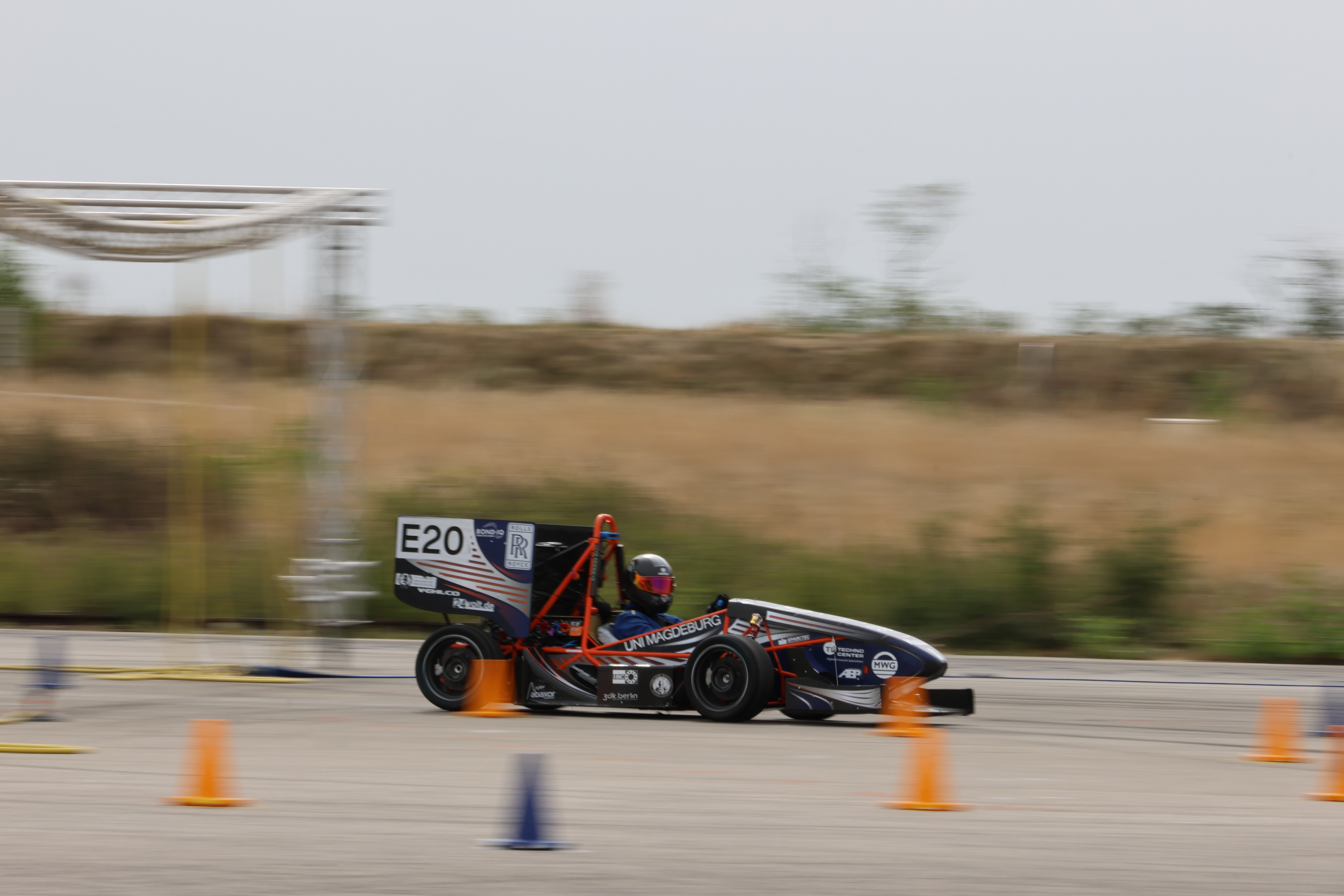 Blau-weiß-oranger Formula Student Rennwagen des UMD Racing e.V. (Universität Magdeburg) mit der Startnummer E20, der schnell auf einer Rennstrecke fährt. Der Rennwagen verwendet CPC Schnellkupplungen vom Schellen-Shop für sein Akkukühlsystem.