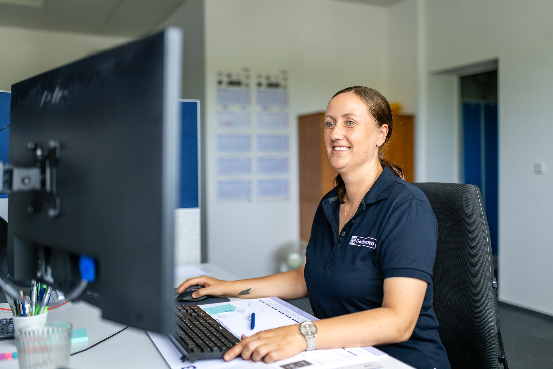 Mitarbeiterin beim Arbeiten am Computer im Büro. Eine lächelnde Frau in einem dunkelblauen Poloshirt mit Firmenlogo sitzt an einem Büroschreibtisch und bedient einen Computer.
